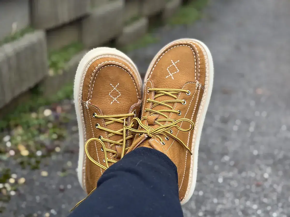 Scarpe marroni in pelle scamosciata, con lacci gialli e cuciture bianche, che sembrano una scarpa a forma di piede.