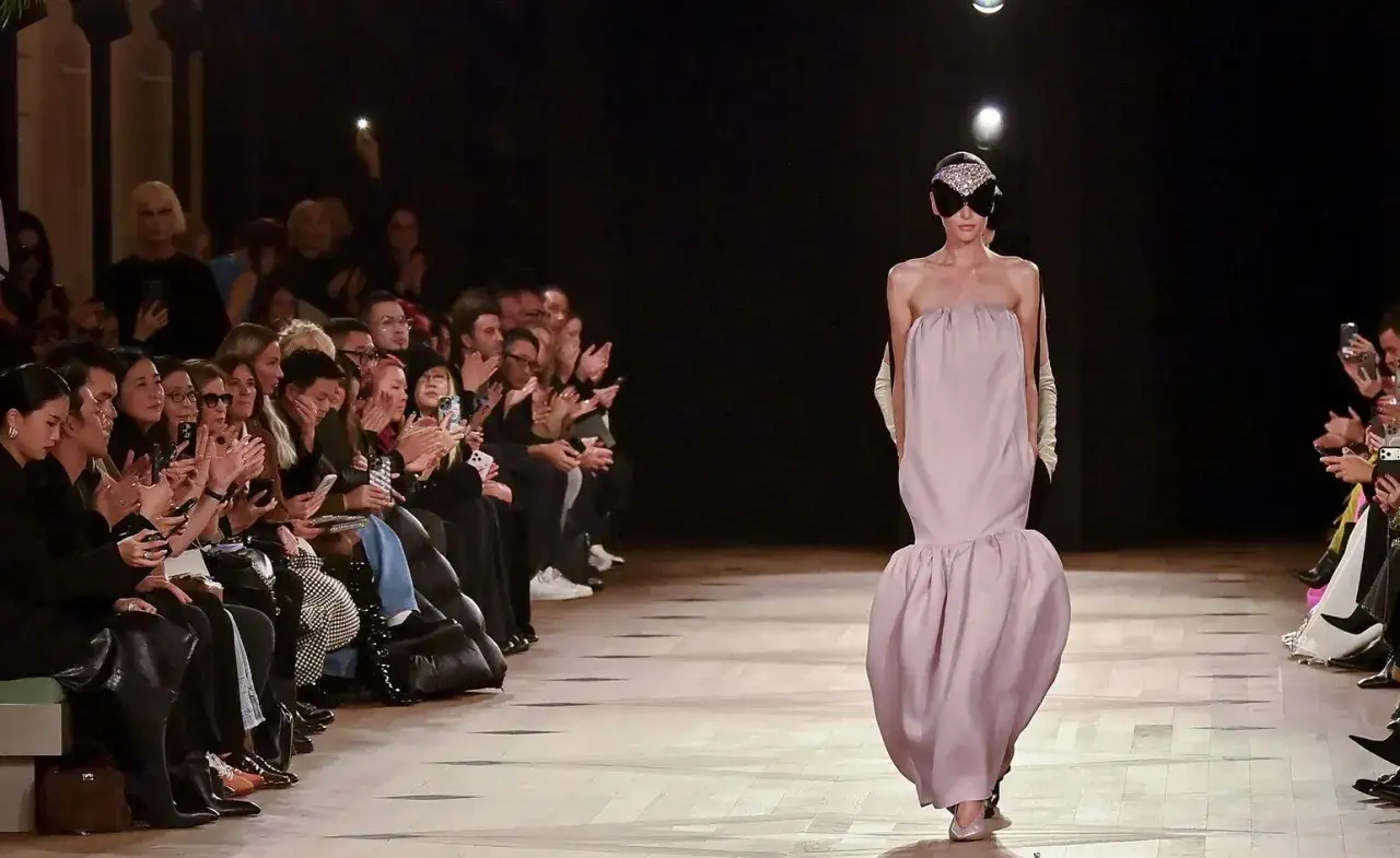 Sfilata di moda: un modello indossa un abito rosa pallido con un'acconciatura audace, mentre il pubblico applaude. L'eleganza di Pierpaolo Piccioli.