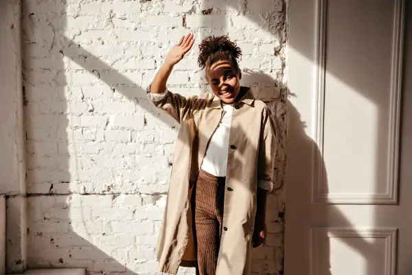 Donna con trench beige e pantaloni marroni, con la mano alzata per proteggersi dal sole. Un outfit versatile, ma quali scarpe abbinare?