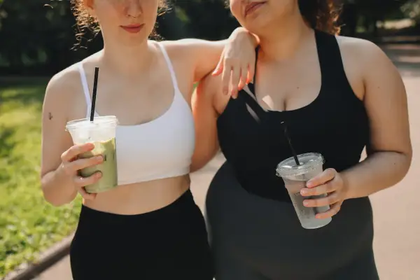 Due amiche con fianchi a violino si godono una bevanda fresca dopo l'allenamento, una con un matcha, l'altra con acqua.