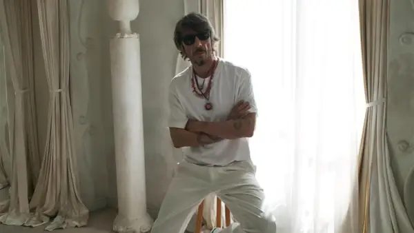 Pierpaolo Piccioli, con occhiali da sole e collane, posa con le braccia conserte in un ambiente chiaro con tende e colonne.