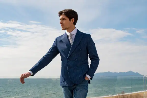 Uomo con giacca blu a doppio petto a righe, camicia bianca e cravatta, appoggiato a una ringhiera con vista mare.