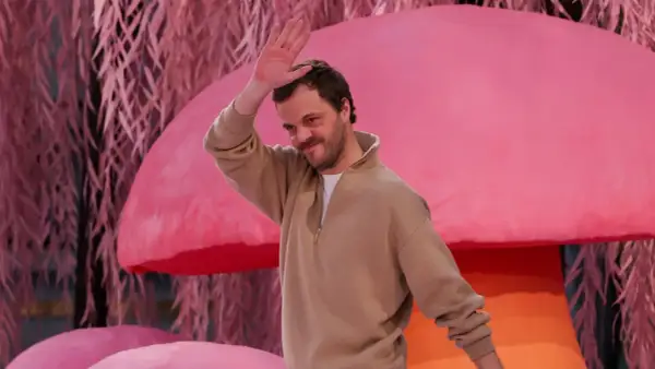 Matthieu Blazy saluta con la mano, sorridendo, su uno sfondo rosa con elementi che ricordano funghi e piante.