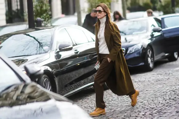 Donna con un elegante outfit con Timberland, cappotto verde oliva, dolcevita bianco e pantaloni marroni, cammina su un acciottolato.