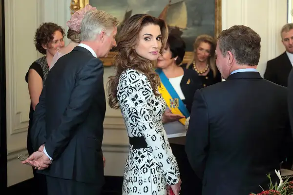 Regina Rania di Giordania, elegante in abito bianco e nero, partecipa a un evento. Sullo sfondo, un quadro e altre personalità.