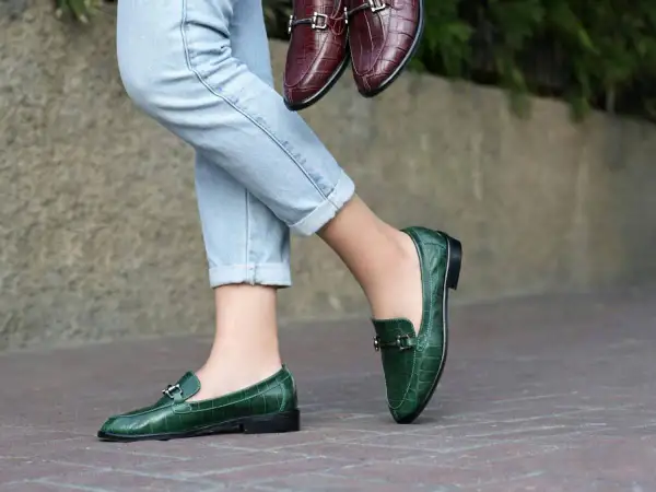 Un paio di jeans chiari e un paio di eleganti mocassini uomo verdi con fibbia metallica. Un altro paio di mocassini marroni è tenuto in mano.