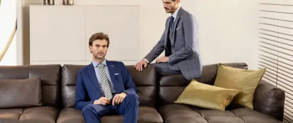 Due uomini eleganti in lounge suit siedono su un divano di pelle marrone. Uno è blu, l'altro grigio a quadri.