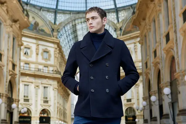 Giovane uomo con un caban blu scuro, un capo che incarna eleganza e calore, passeggia sotto la volta di una galleria storica.