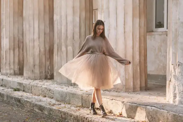 Ragazza con un outfit gonna tulle lunga color cipria, maglione oversize e anfibi, passeggia tra colonne antiche.