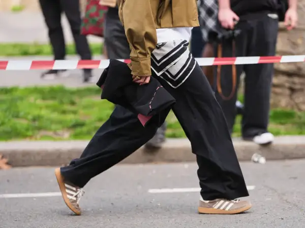 Persona con un outfit casual, pantaloni neri ampi, giacca color kaki e scarpe da ginnastica Adidas marroni.