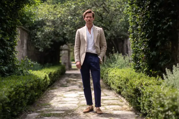 Uomo con giacca beige e pantaloni blu, un look che incarna lo stile francese uomo, in un giardino lussureggiante.