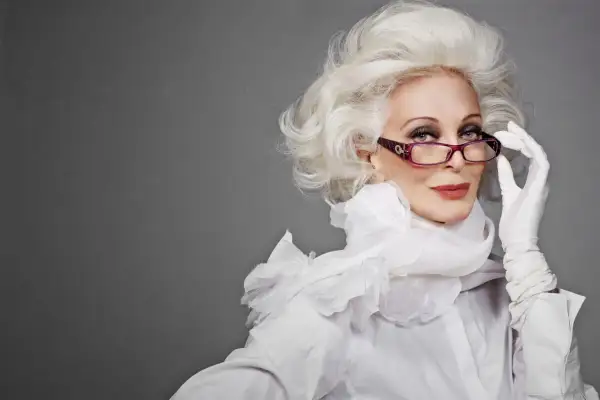 Carmen Dell'Orefice, icona di stile, con capelli bianchi voluminosi, occhiali viola e guanti bianchi, posa con eleganza.