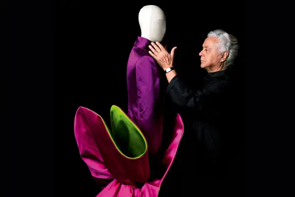 Roberto Capucci, maestro della moda, accarezza un abito viola su un manichino, circondato da petali fucsia e verdi.