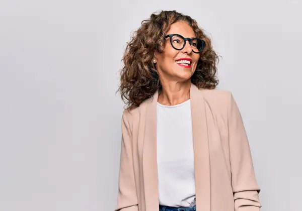Donna sorridente con occhiali e blazer beige, un esempio di come vestirsi a 50 anni con stile.