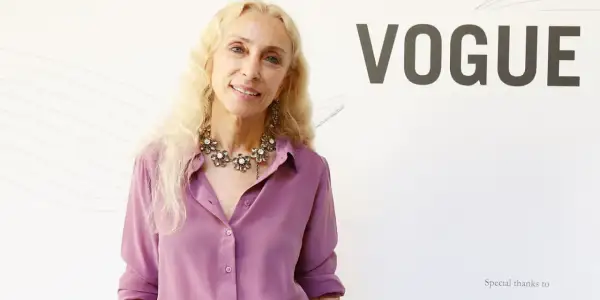 Franca Sozzani, icona di stile, sorride con un collier floreale e una camicia lilla, sullo sfondo del logo VOGUE.
