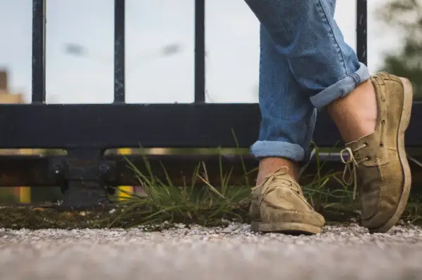 Gambe con jeans arrotolati e scarpe senza calzini, appoggiate su un muretto con erba e ghiaia.