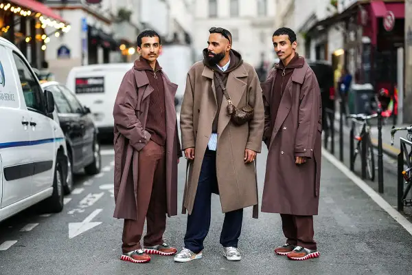 Tre uomini con eleganti stili abbigliamento uomo, cappotti lunghi e pantaloni coordinati, posano in strada.
