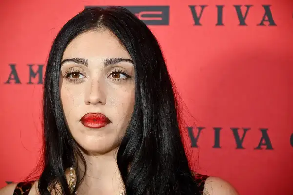 Lourdes Leon, con labbra rosse audaci e sguardo intenso, posa davanti a uno sfondo rosso con scritte nere.